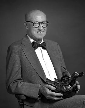 Retrato en blanco y negro de George M. Foster, sentado, con gafas, pajarita y traje, sosteniendo una escultura antropológica en sus manos.